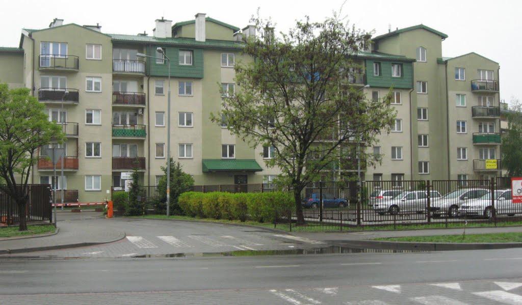 Wielopiętrowy budynek mieszkalny z balkonami, ogrodzony metalowym płotem i szlabanem, widoczny z mokrej ulicy po deszczu.