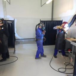 Oliweld - Trzech pracowników w kombinezonach i maskach ochronnych, pracujących w warsztacie spawalniczym, iskrzące się spoiny, stanowiska oddzielone czarnymi kurtynami.