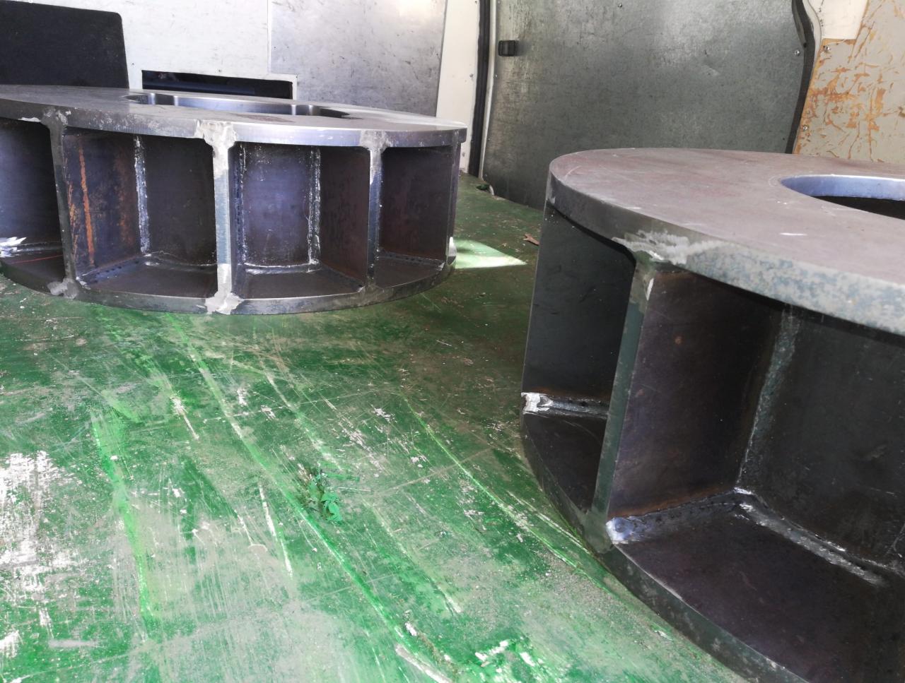 Dwa duże, metalowe elementy konstrukcyjne z widocznymi spawami, leżące na zielonej, porysowanej podłodze, prawdopodobnie w hali produkcyjnej.