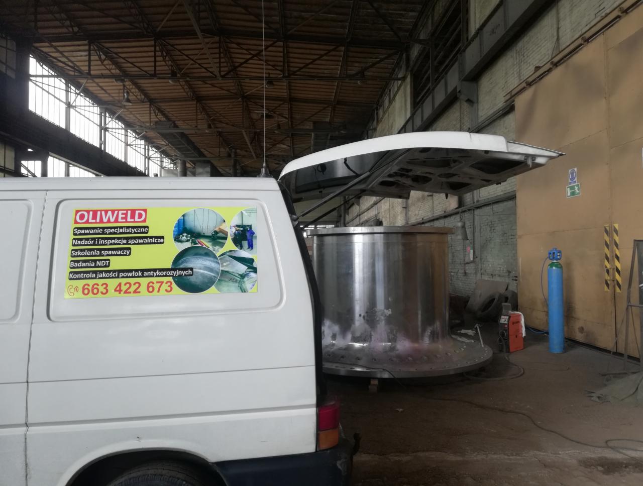 Cylindryczny metalowy zbiornik w hali produkcyjnej, widoczny obok biały samochód dostawczy z naklejką firmy OLIWELD oferującej spawanie specjalistyczne, nadzór spawalniczy i badania NDT.