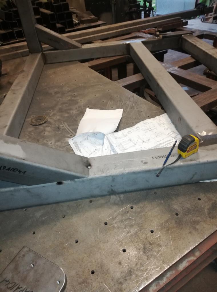 Metalowa konstrukcja ramowa w trakcie tworzenia, leżąca na stole warsztatowym, obok rysunek techniczny, ołówek i miarka zwijana.