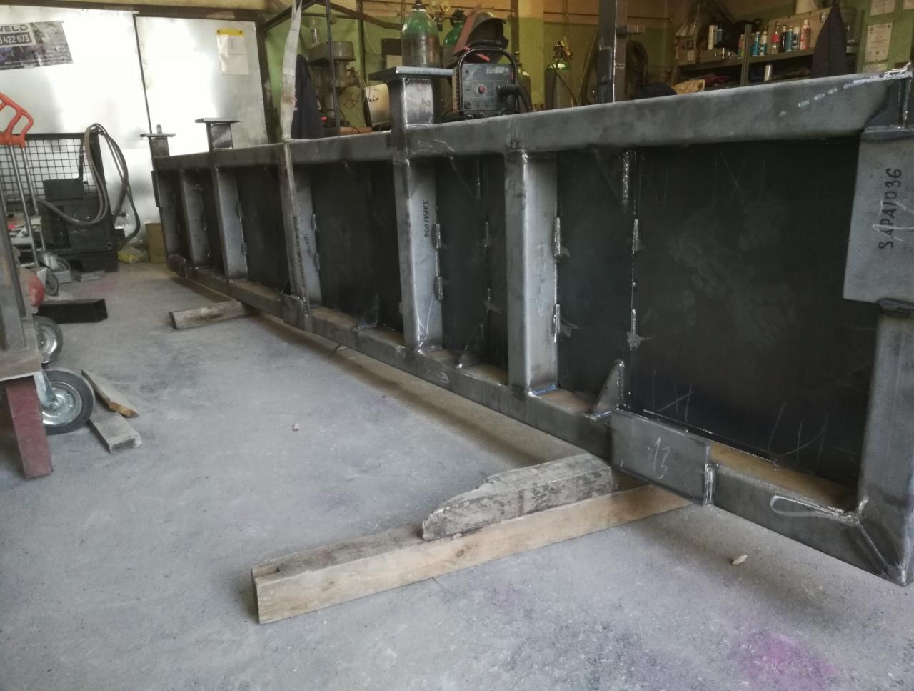 Spawana konstrukcja stalowa z profili zamkniętych, podparta drewnianymi klockami, widoczna w warsztacie.