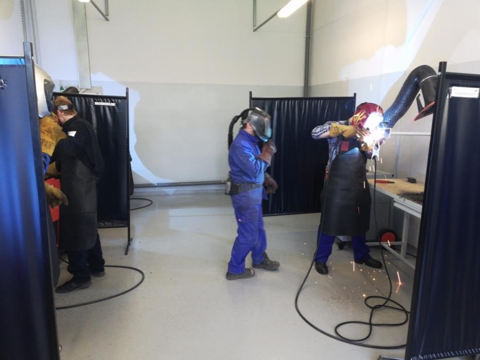 Trzech pracowników w kombinezonach i maskach ochronnych, pracujących w warsztacie spawalniczym, iskrzące się spoiny, stanowiska oddzielone czarnymi kurtynami.