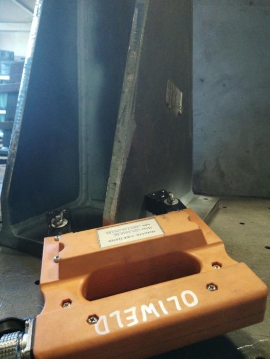 Kontrola spoiny magnetycznym jarzmem OLIWELD na metalowej konstrukcji spawanej, widoczny przewód zasilający i naklejka informacyjna na urządzeniu.