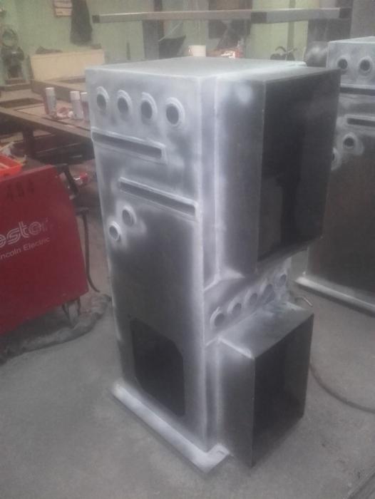 Metalowa obudowa z otworami i prostokątnymi wnękami, świeżo po malowaniu, widoczny czerwony sprzęt spawalniczy w tle.