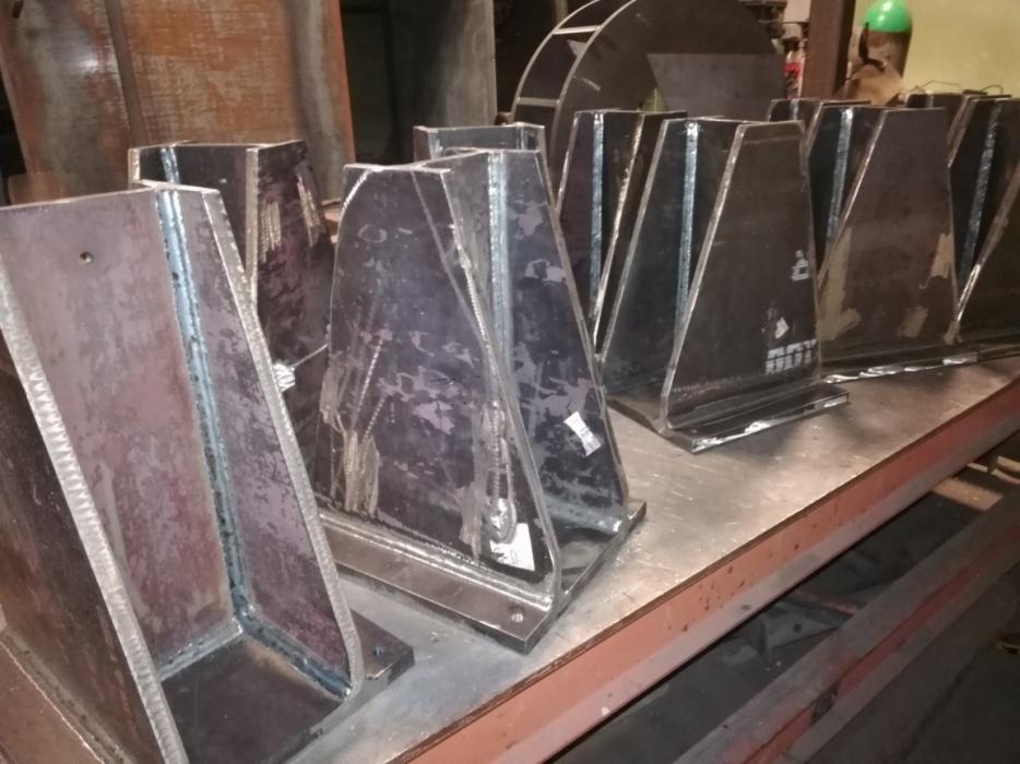 Spawane, metalowe elementy konstrukcyjne w kształcie trapezów, ułożone na stole warsztatowym, widoczne ślady spawania i obróbki metalu.