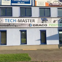 Budynek firmy Tech-Master z banerem reklamującym agregaty malarskie, szpachlarskie i tynkarskie marek Graco i Festool, widoczne okna w niebieskich ramach i chodnik z kostki brukowej.