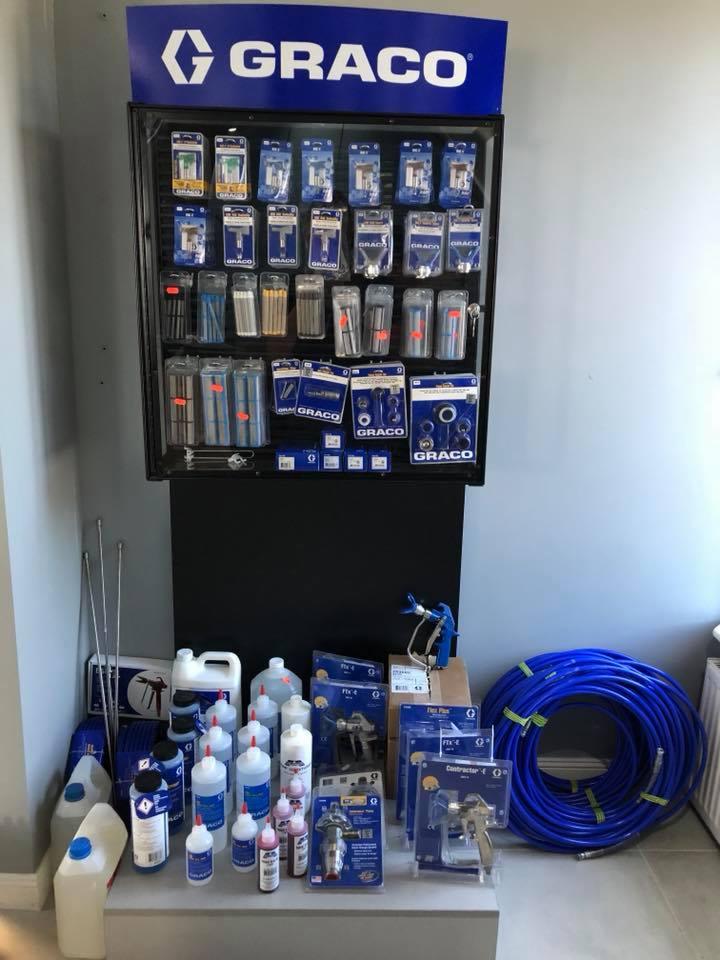 Ekspozycja produktów marki Graco: dysze, filtry, pistolety natryskowe w opakowaniach, pojemniki z płynami i zwój niebieskiego węża ciśnieniowego.