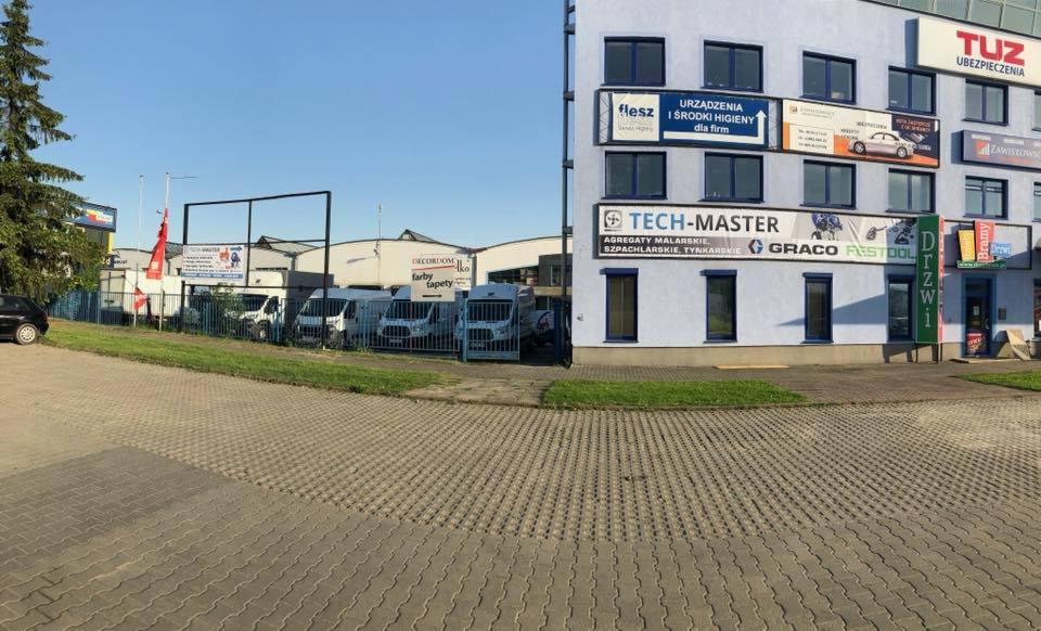 Widok na budynek firmy z reklamami na elewacji, zaparkowane busy za ogrodzeniem, oraz fragment parkingu z kostki brukowej.