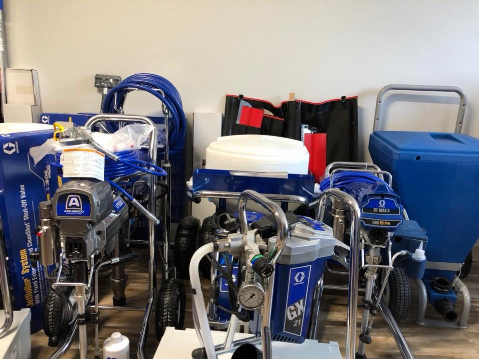 Zestaw profesjonalnych agregatów malarskich różnych modeli, w odcieniach szarości i niebieskiego, z widocznymi wężami i akcesoriami, prezentowany w pomieszczeniu.