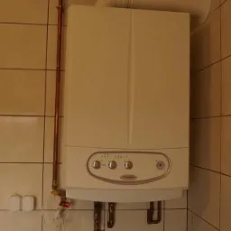 Biały piec gazowy zamontowany na ścianie z beżowych płytek, z widocznymi miedzianymi rurami doprowadzającymi gaz i odprowadzającymi spaliny.