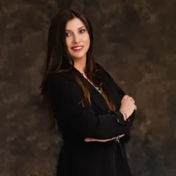 Portret uśmiechniętej adwokatki w czarnej marynarce, z rękami skrzyżowanymi na tle abstrakcyjnego, ciemnego tła.