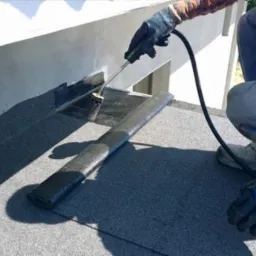 Pracownik w rękawicach aplikuje uszczelniacz pędzlem z przewodem na płaski dach pokryty papą, widoczny fragment ściany budynku.