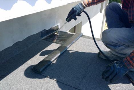 Pracownik w rękawicach aplikuje uszczelniacz pędzlem z przewodem na płaski dach pokryty papą, widoczny fragment ściany budynku.