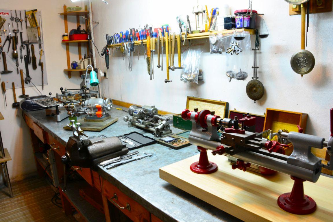 Warsztat z różnymi tokarkami na stole warsztatowym, narzędzia wiszące na ścianie, w tle widać wagę szalkową i zegar z wahadłem.