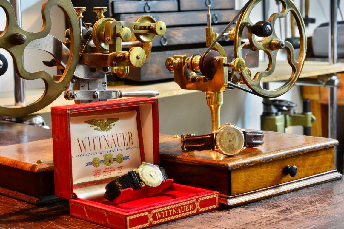 Warsztat zegarmistrzowski: Dwa zabytkowe zegarki Wittnauer, jeden w czerwonym pudełku z logo, drugi obok, na tle precyzyjnych narzędzi zegarmistrzowskich z kołami zamachowymi.