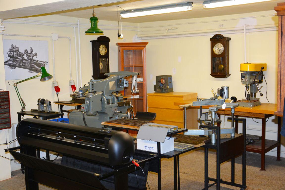 Warsztat z różnymi maszynami: tokarka, wiertarka, ploter tnący, oraz narzędzia i akcesoria. Na ścianach wiszą zegary i plakat z rysunkiem technicznym tokarki.