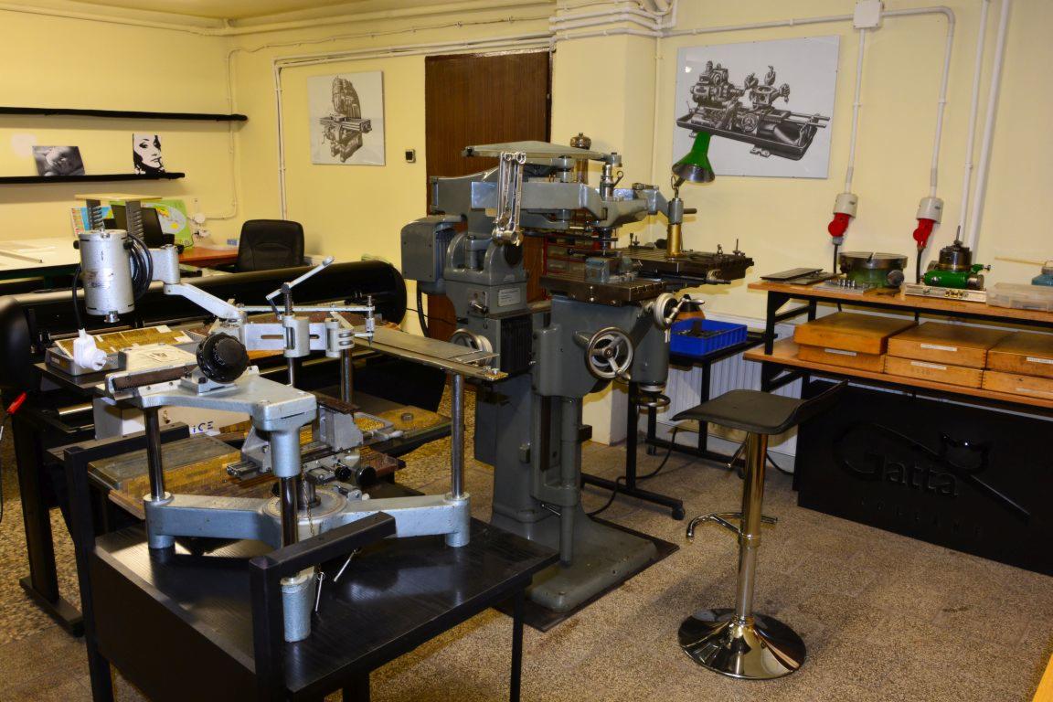 Warsztatowa przestrzeń z frezarką pionową, stolikiem pomocniczym i chromowanym krzesłem barowym na tle żółtych ścian i rysunków technicznych.