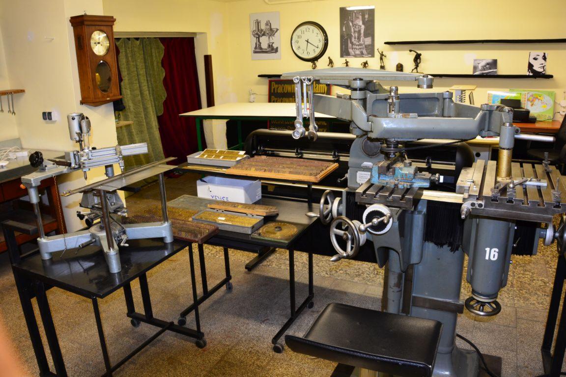 Warsztat z frezarką metalu, stołem z czcionkami drukarskimi i narzędziami w tle.