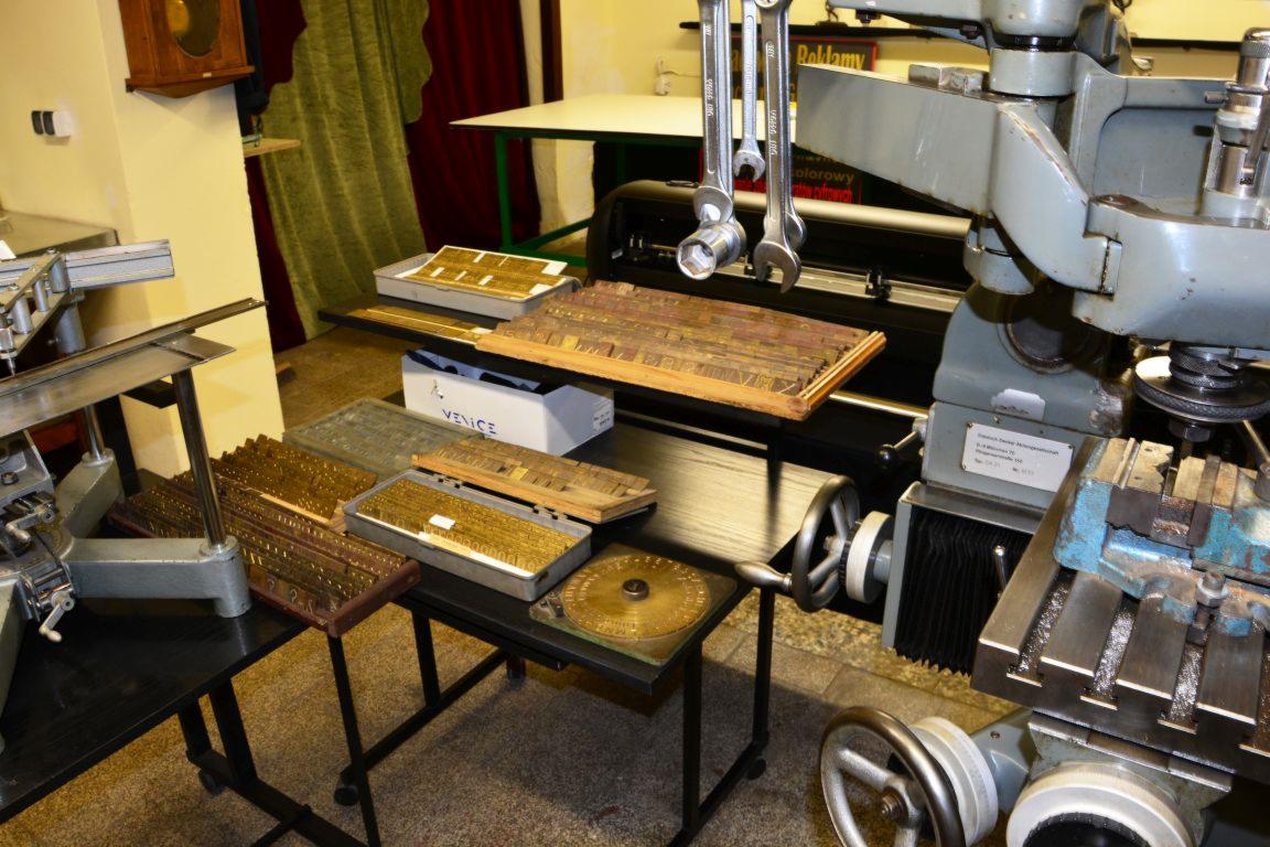 Warsztatowa sceneria z metalowymi czcionkami drukarskimi w kasetach, kluczami nasadowymi wiszącymi nad maszyną drukarską oraz fragmentem frezarki w tle.