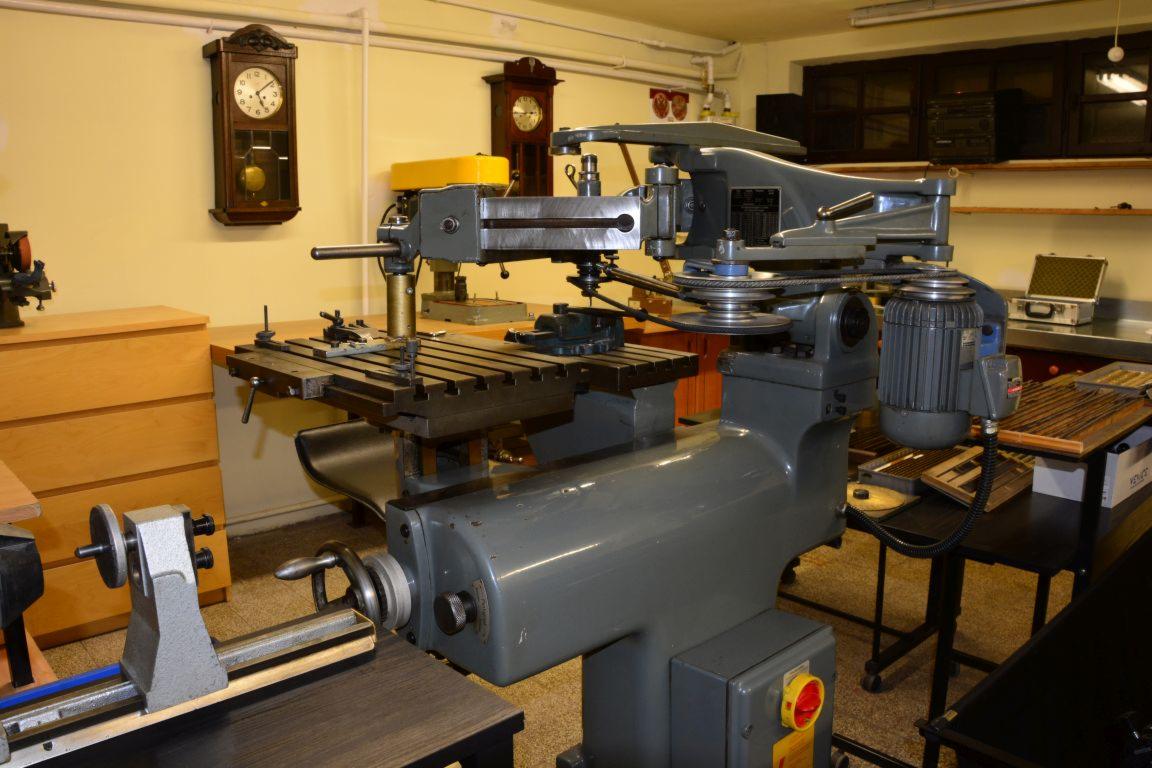 Szara frezarka uniwersalna z metalowym stołem roboczym, widoczne pokrętła, silnik elektryczny i pasek klinowy, w tle warsztat z zegarami i szafkami.