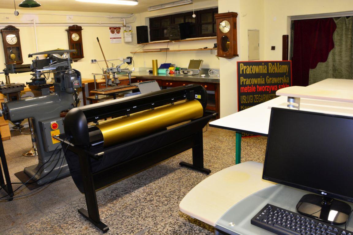 Szeroki ploter tnący z rolką złotej folii w warsztacie, obok tokarka i biurko z komputerem, w tle tablica informacyjna 'Pracownia Reklamy'.