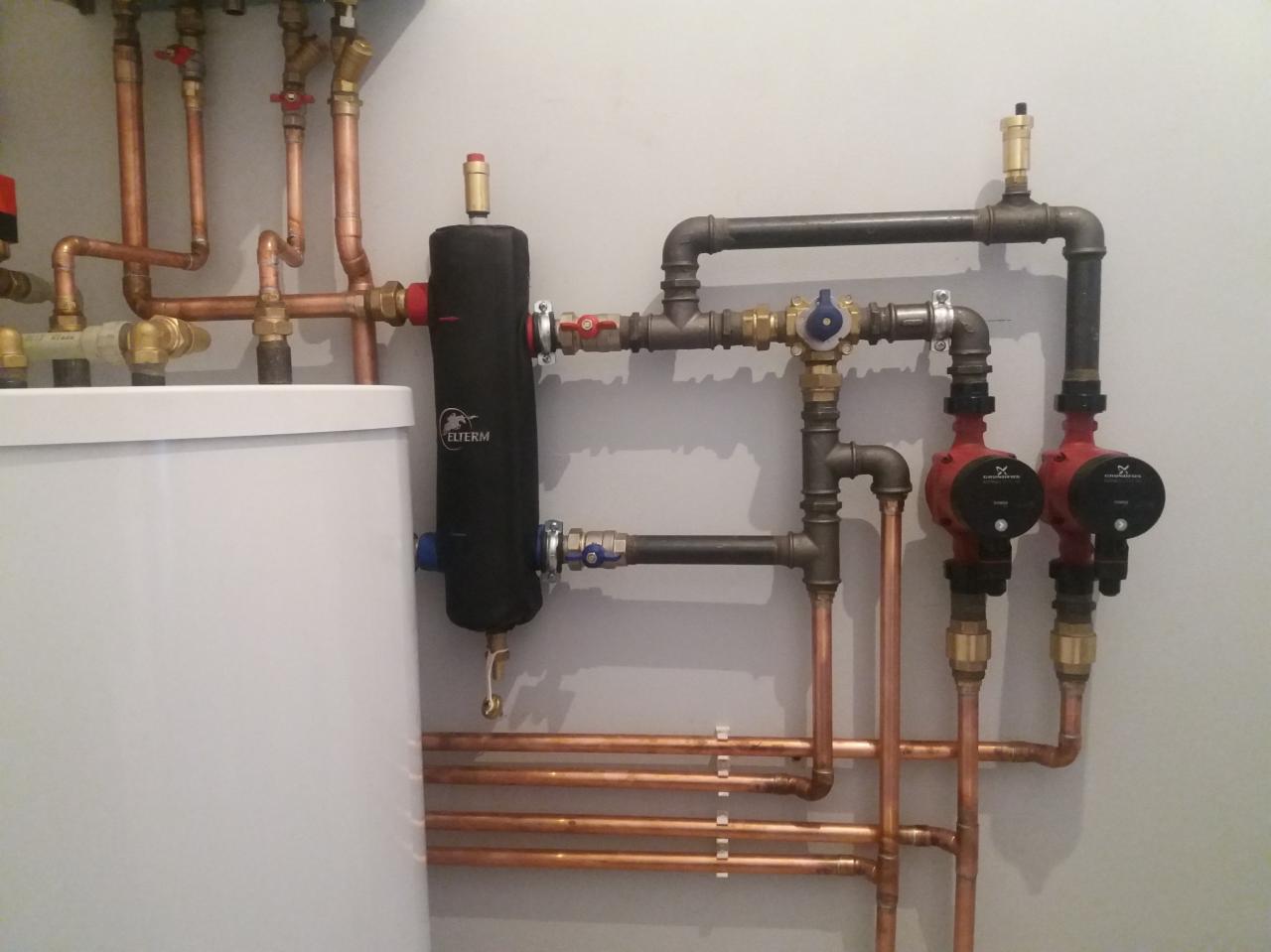 Instalacja centralnego ogrzewania z kotłem, czarnym filtrem Elterm, miedzianymi rurami, zaworami i dwoma czerwonymi pompami Grundfos na szarej ścianie.