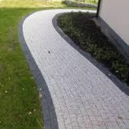 Zakrzywiony chodnik z jasnoszarej kostki brukowej, obramowany ciemniejszą kostką, prowadzący wzdłuż budynku. Widoczny fragment trawnika i rabaty z ziemią.