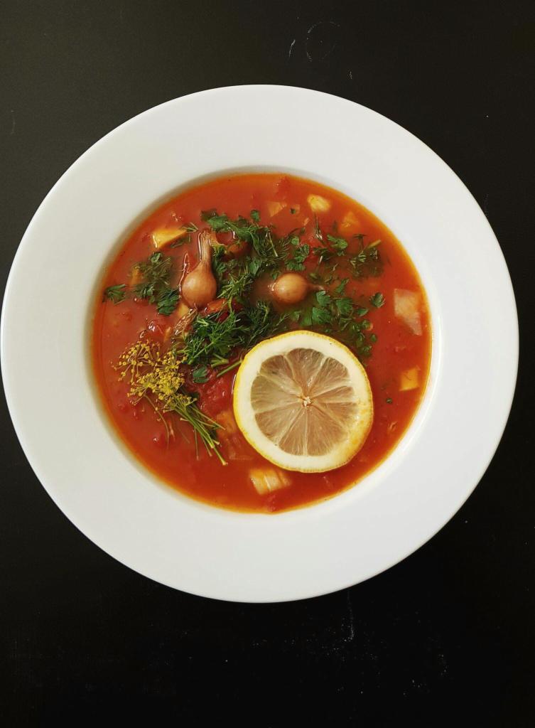 Apetyczna zupa pomidorowa w białym talerzu, udekorowana plastrem cytryny, świeżym koperkiem i młodymi cebulkami, na czarnym tle.