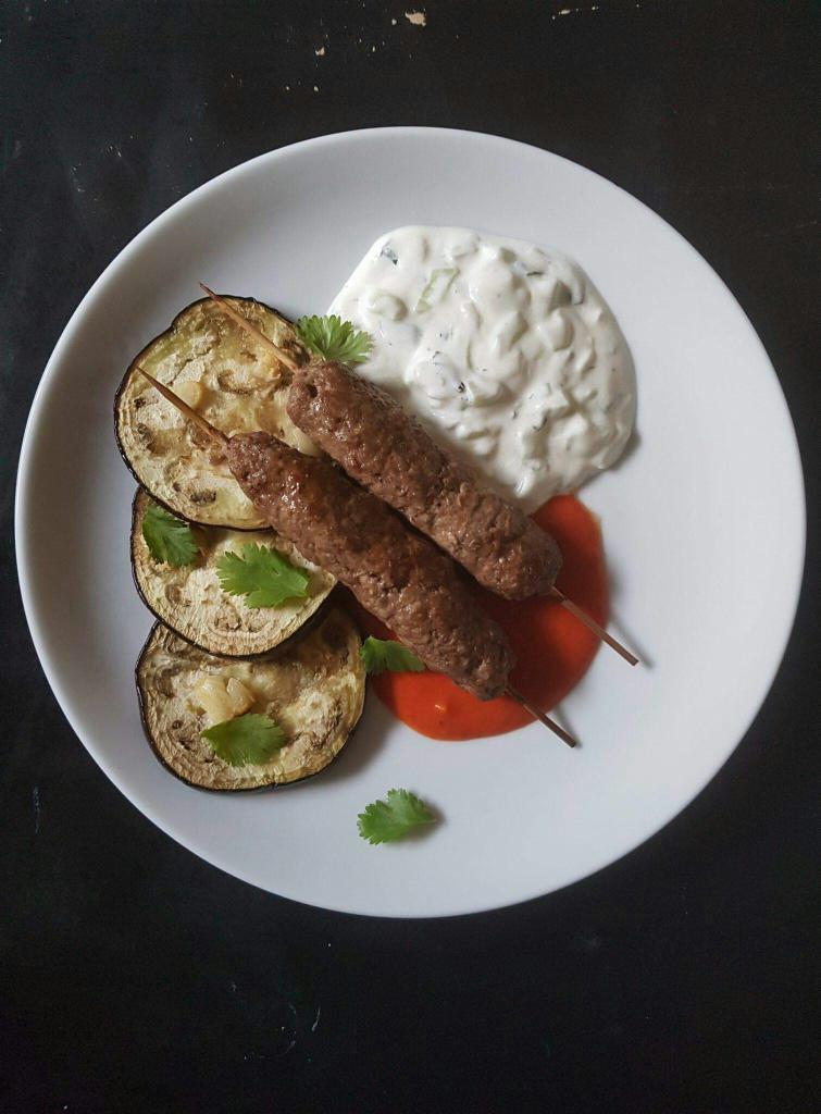 Dwa szaszłyki z mięsa mielonego na drewnianych patyczkach, podane na białym talerzu z grillowanym bakłażanem, sosem tzatziki i sosem pomidorowym, udekorowane świeżą kolendrą.
