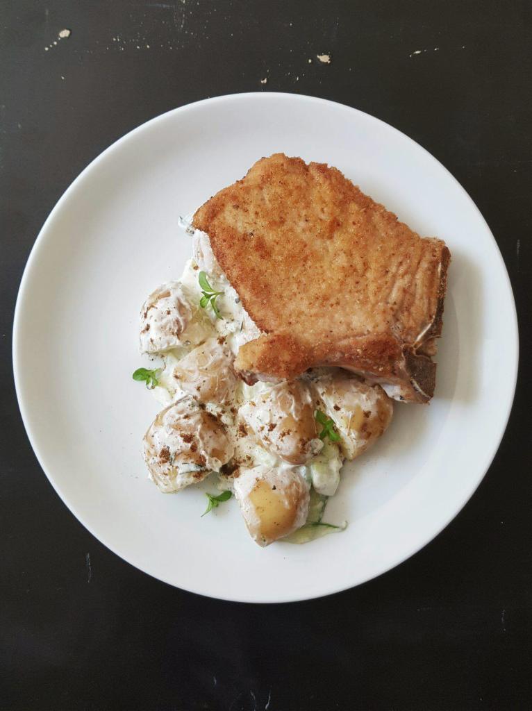 Kotlet schabowy z kością i sałatka ziemniaczana z ziołami na białym talerzu, czarne tło.