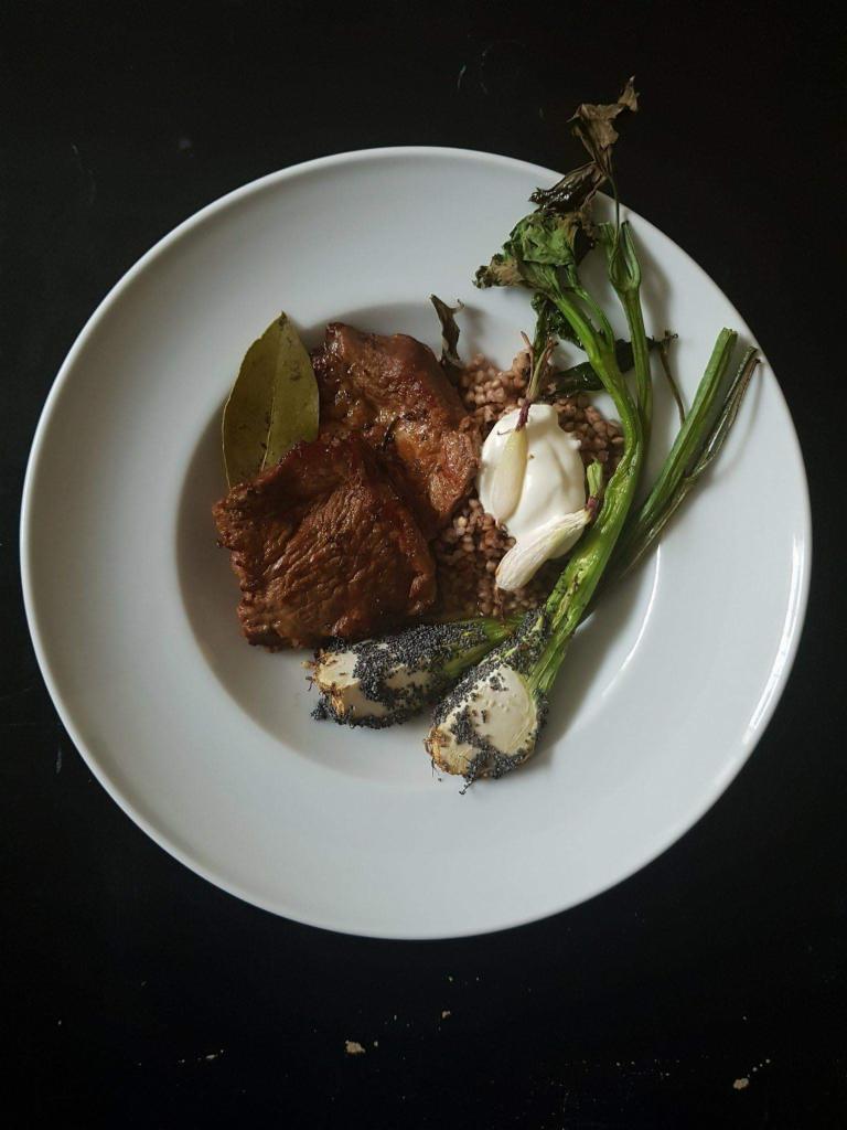 Porcja duszonej wołowiny z kaszą gryczaną, pieczonym czosnkiem i cebulką dymką na białym talerzu, ujęcie z góry na czarnym tle.