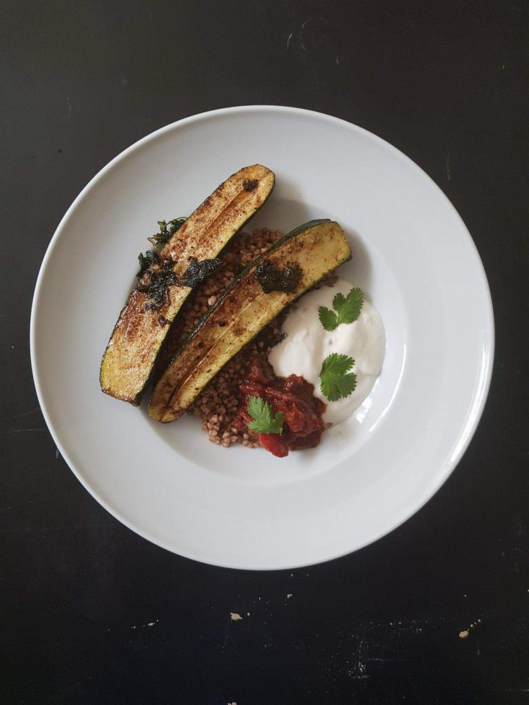 Dwa grillowane plastry cukinii na kaszy gryczanej z sosem pomidorowym i kleksem jogurtu, udekorowane świeżą kolendrą na białym talerzu na ciemnym tle.