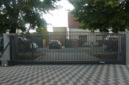 Nowoczesna, przesuwna brama wjazdowa z pionowych, metalowych profili, szara kolorystyka, widoczny napęd elektryczny, kostka brukowa na podjeździe, dom jednorodzinny w tle.