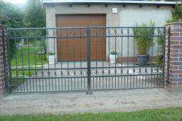 Czarna, metalowa brama wjazdowa z dekoracyjnymi okręgami, osadzona między dwoma słupkami z czerwonej cegły. W tle widoczny garaż z brązową bramą roletową i donice z zielonymi roślinami.