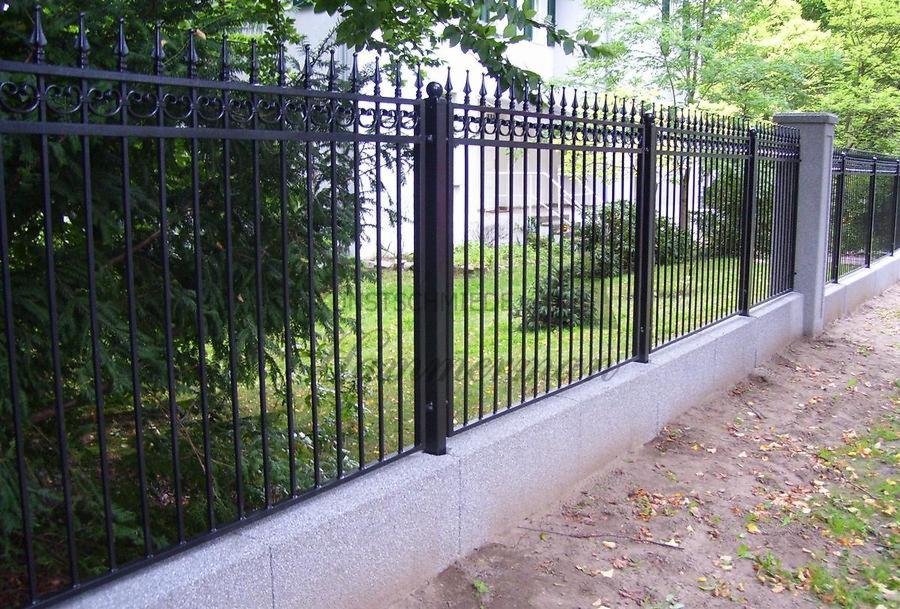Czarne, metalowe ogrodzenie z kutymi zdobieniami na szczytach prętów, osadzone na niskiej, szarej podmurówce z widocznymi kamyczkami, z zielonymi krzewami i drzewami w tle.