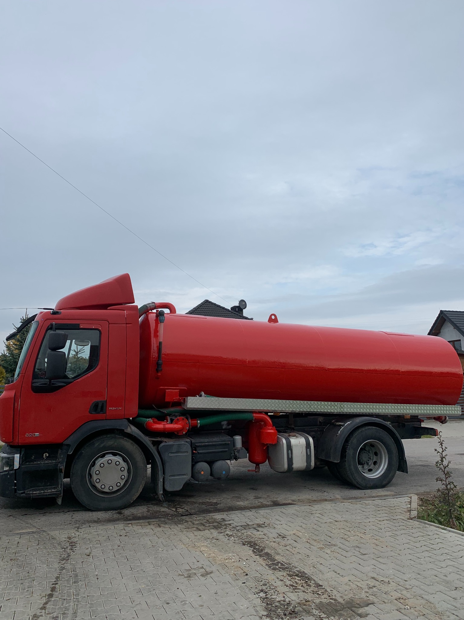 Czerwony samochód asenizacyjny z błyszczącym zbiornikiem na podwoziu ciężarówki, zaparkowany na brukowanej powierzchni, z widocznymi elementami instalacji i zielonymi wężami.
