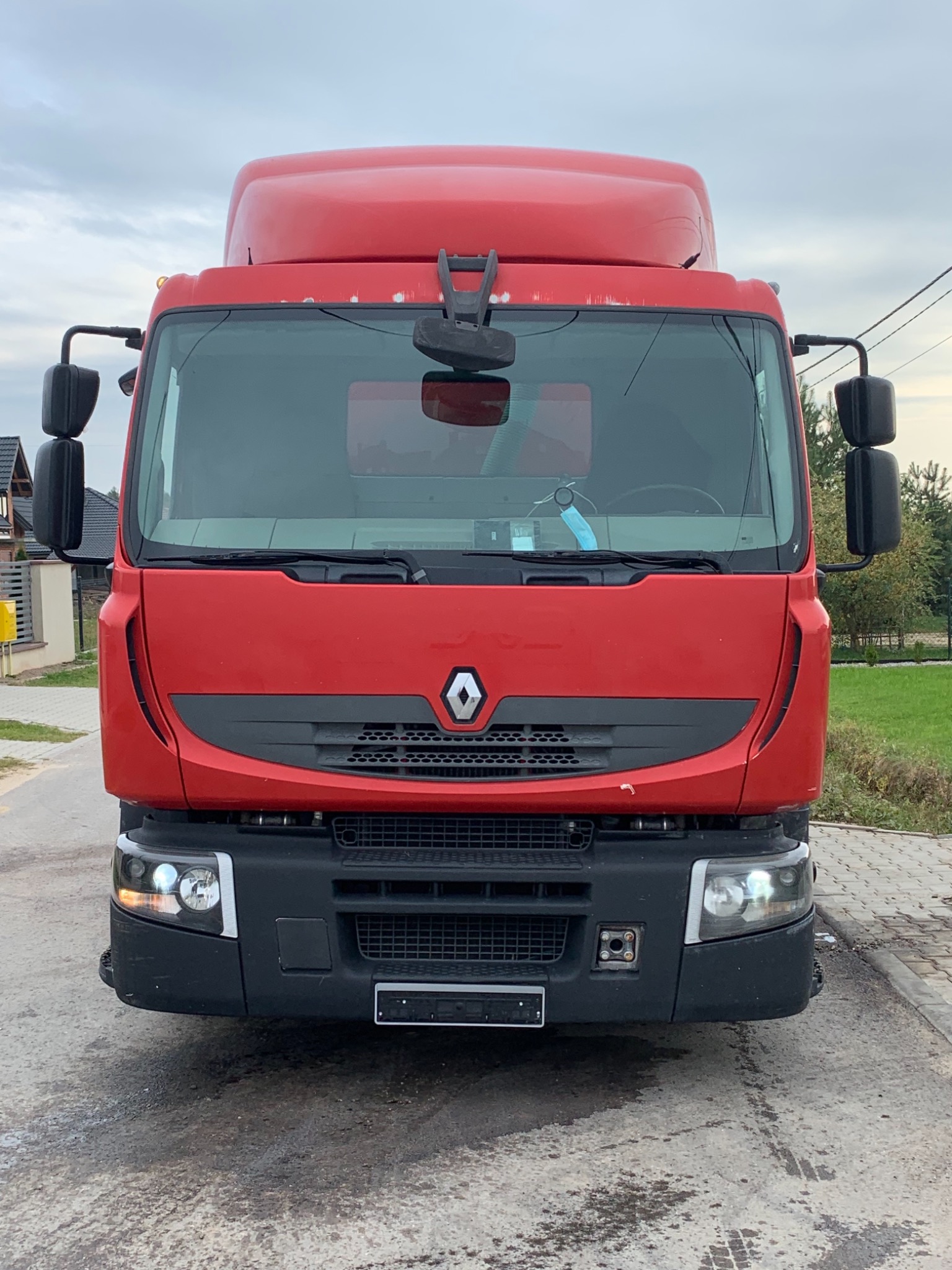 Czerwony samochód ciężarowy Renault z widoczną maską i reflektorami, zaparkowany na betonowej nawierzchni, z maską chirurgiczną wiszącą na lusterku wstecznym.