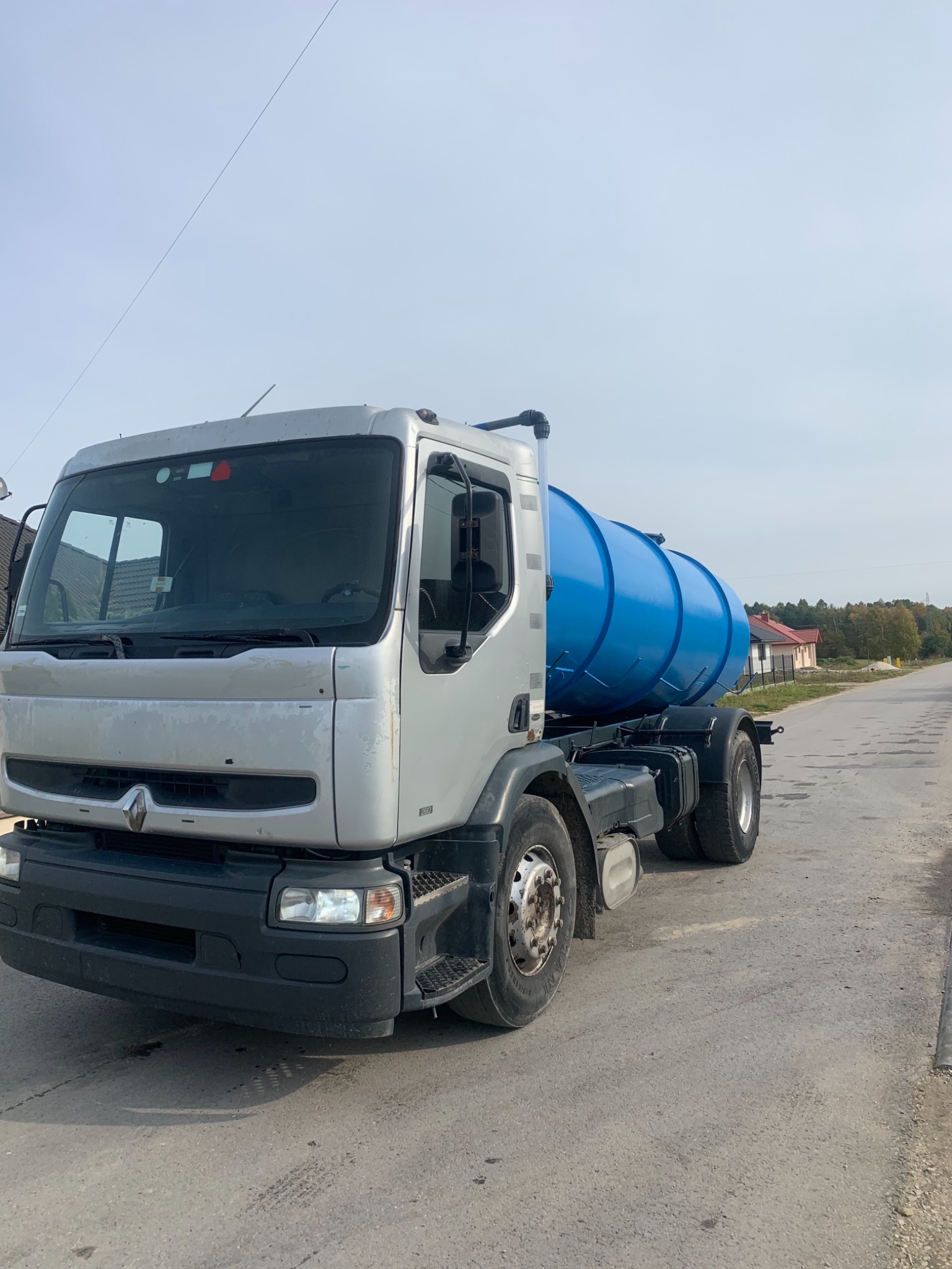 Srebrna ciężarówka Renault z niebieskim zbiornikiem cysterną na podwoziu, stojąca na asfaltowej drodze w słoneczny dzień.