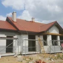 Dom w trakcie budowy z czerwoną dachówką, widoczne rusztowania i pozostałości materiałów budowlanych na placu.