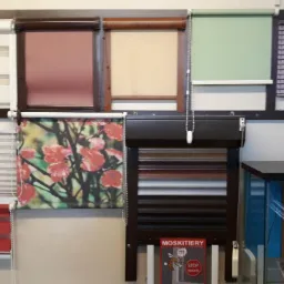 Ekspozycja różnych typów osłon okiennych: rolety dzień i noc, z nadrukiem kwiatowym, żaluzje drewniane w odcieniach brązu, oraz reklama moskitier w salonie.