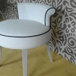 Tapicerowany stołek w stylu glamour, biała pikowana skóra ekologiczna z czarną lamówką, na tle ściany w ornamenty.