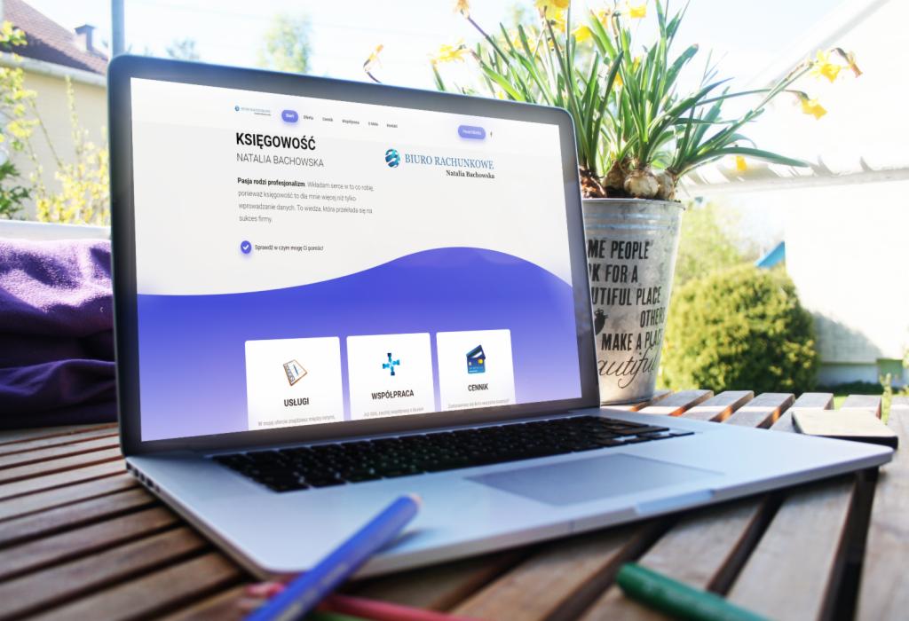 Laptop wyświetlający stronę internetową biura rachunkowego Natalia Bachowska, na drewnianym stole, obok doniczki z narcyzami i kolorowych kredek.