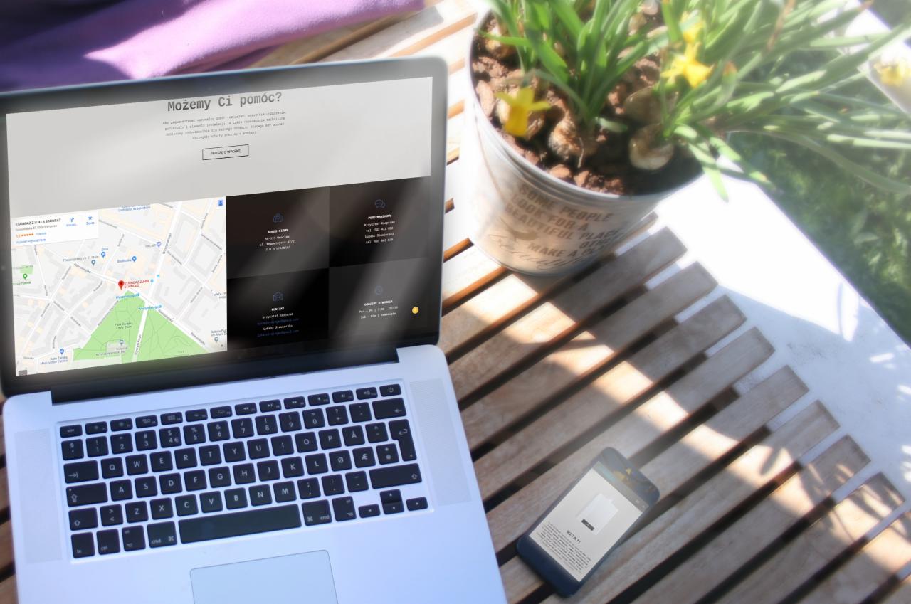 Laptop wyświetlający stronę internetową z mapą i danymi kontaktowymi firmy, obok doniczka z żółtymi kwiatami i smartfon na drewnianym blacie w słoneczny dzień.
