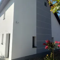 Dwukondygnacyjny dom po malowaniu elewacji, z szarą wstawką i białymi liniami. Widoczna kamera na ścianie, krzew z różowymi kwiatami i elementy ogrodu.