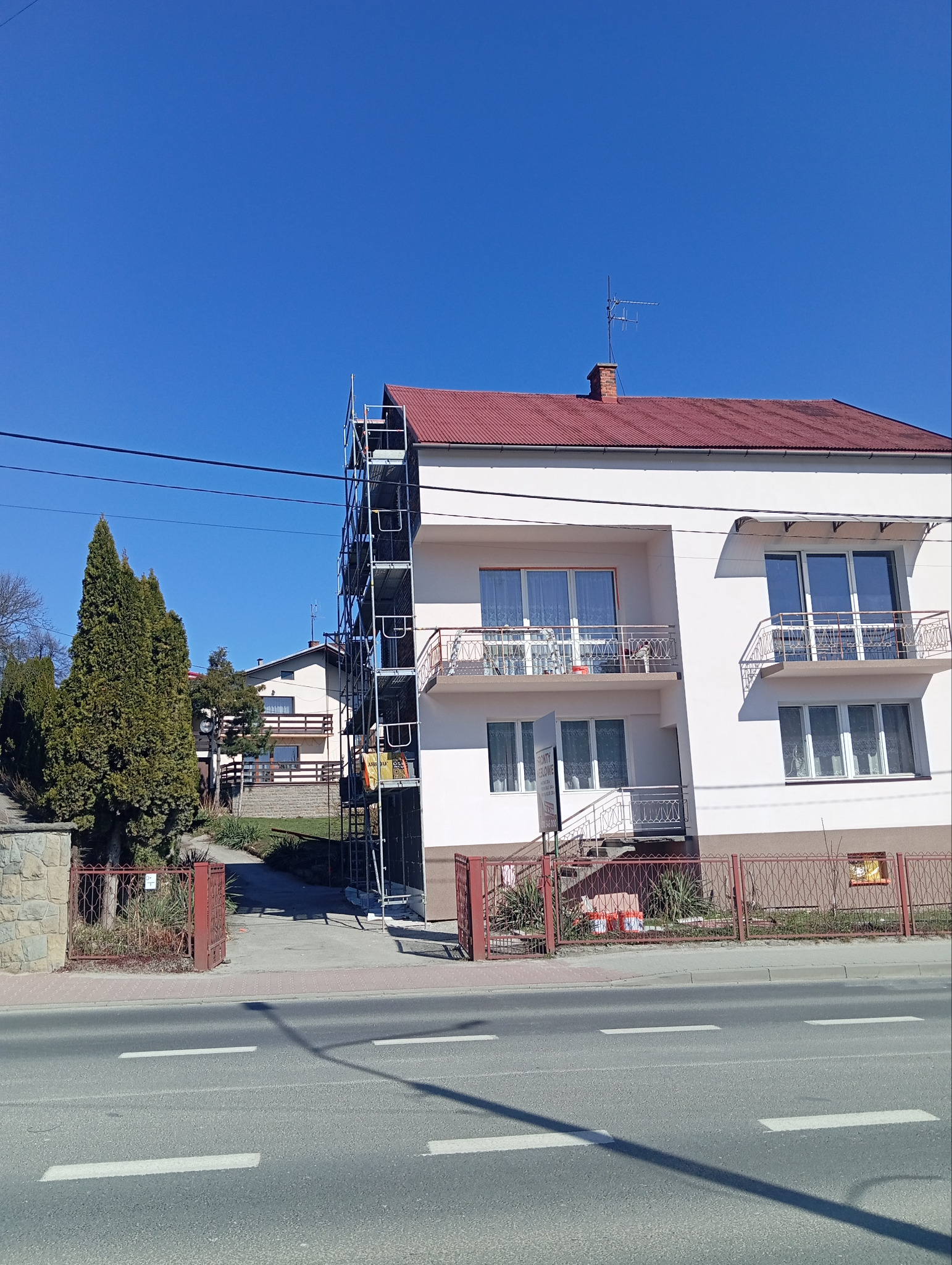 Dwupiętrowy dom z rusztowaniem elewacyjnym, czerwony dach, jasna elewacja. Widok z ulicy, słoneczny dzień. Częściowo widoczny baner reklamowy.
