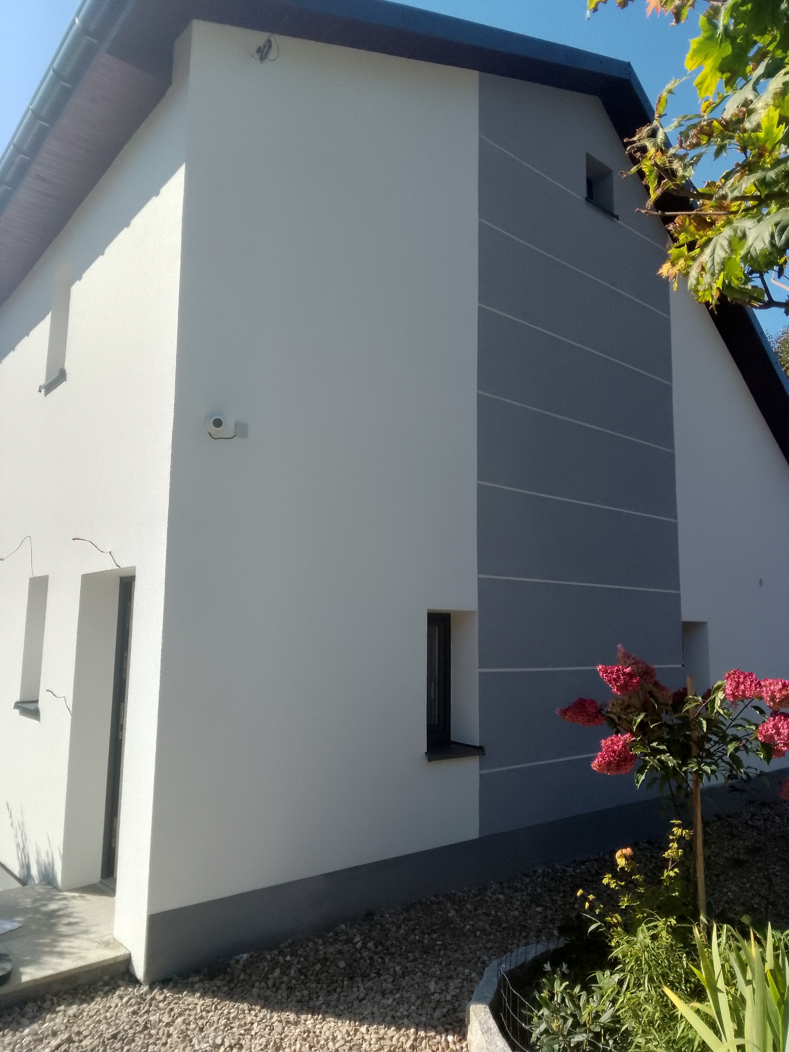 Dwukondygnacyjny dom po malowaniu elewacji, z szarą wstawką i białymi liniami. Widoczna kamera na ścianie, krzew z różowymi kwiatami i elementy ogrodu.