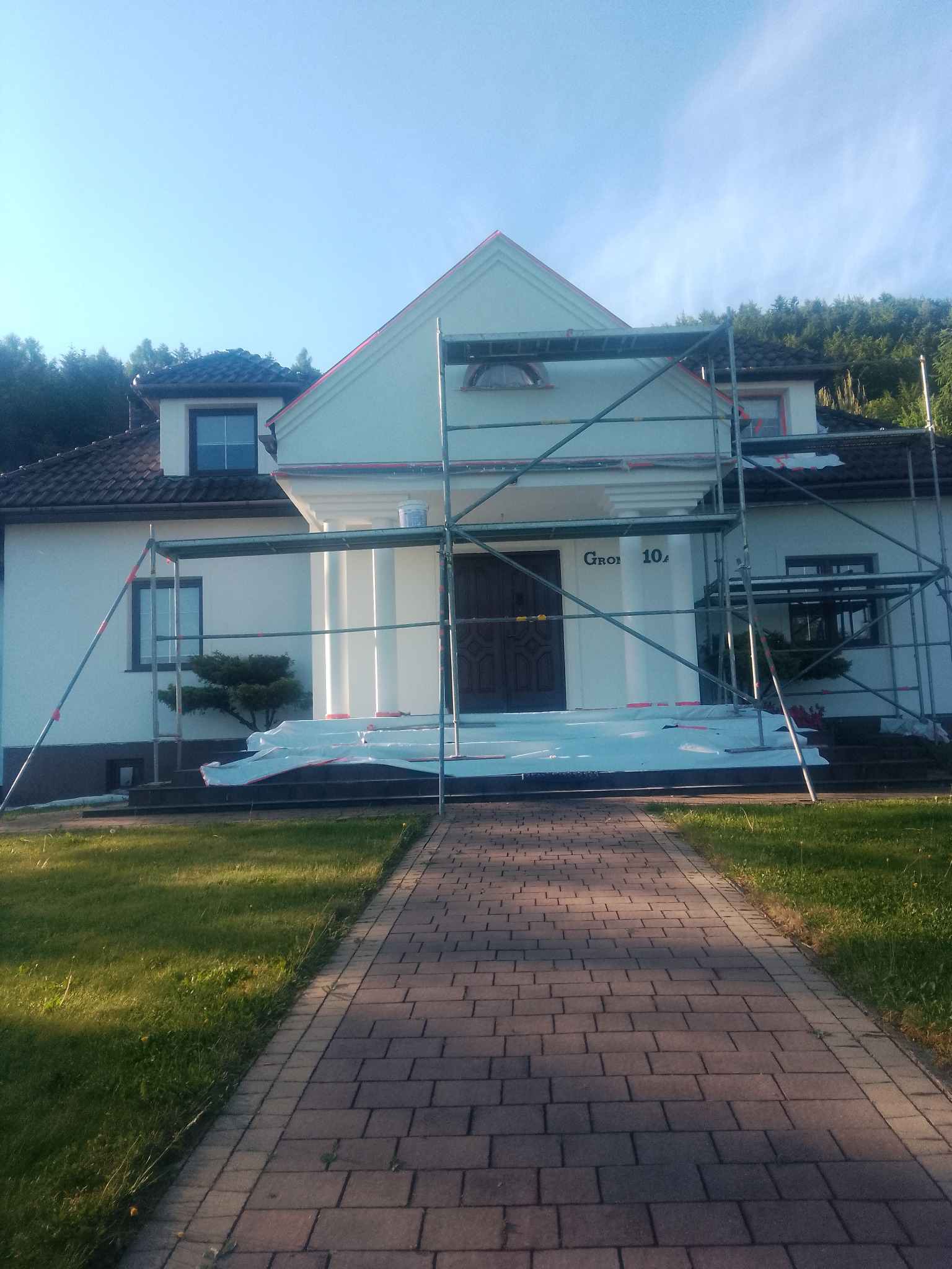 Dom jednorodzinny w trakcie prac elewacyjnych, widoczne rusztowania i zabezpieczenia. Świeżo pomalowana fasada, jasne kolory, schludny wygląd.