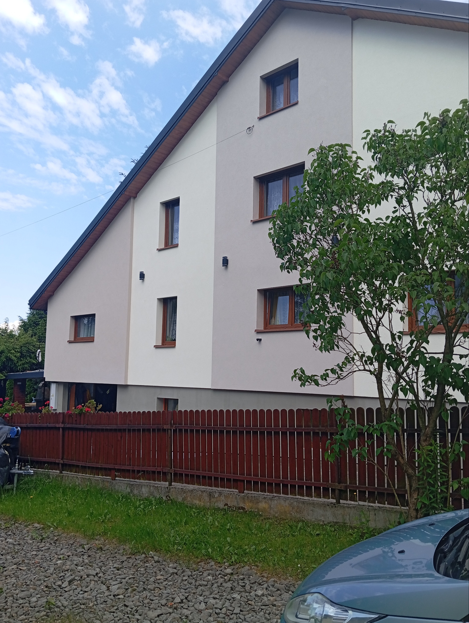 Dwukolorowa elewacja domu jednorodzinnego z brązowymi oknami, widziana z poziomu gruntu, częściowo zasłonięta przez drzewo i ogrodzenie.