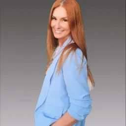 Uśmiechnięta kobieta z rudymi włosami w błękitnej marynarce, pozująca na szarym tle.